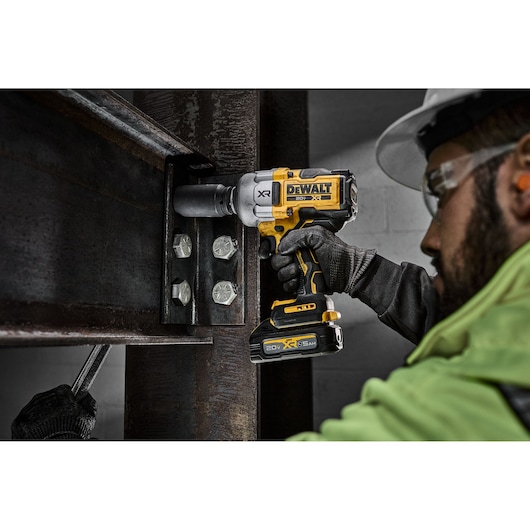 A person using a DEWALT cordless impact wrench (model DCF964B) to tighten bolts on a large steel structure. The worker is wearing gloves and safety gear. The DEWALT tool is prominently featured.