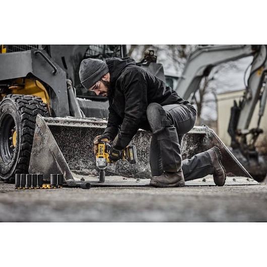 A person wearing outdoor work clothing is kneeling in front of heavy machinery and using a DEWALT cordless impact wrench. Several socket attachments are placed nearby on the ground.