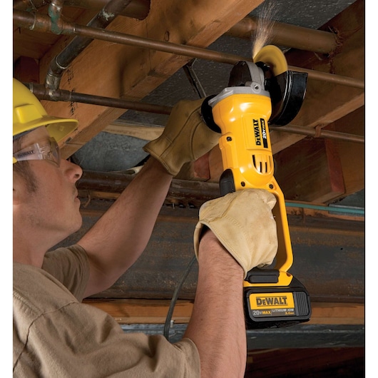 A person wearing work gloves is using a Dewalt DCG412B 20V Max Lithium Ion cordless angle grinder to cut through a metal pipe. The scene is set in a construction or maintenance area with wooden beams and pipes overhead.
