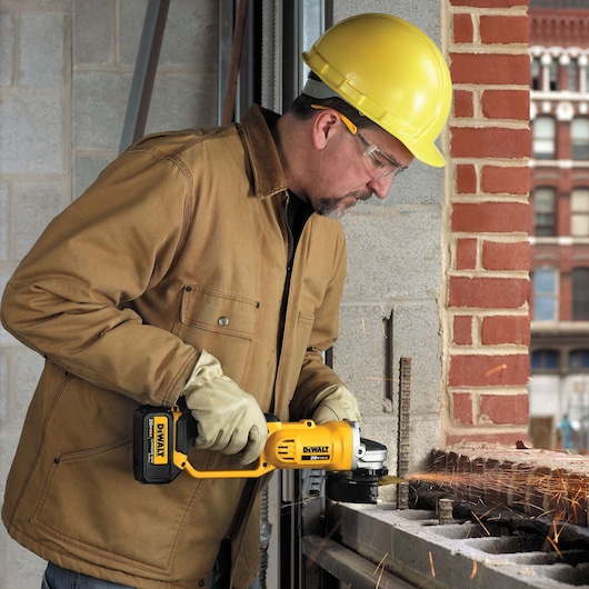 A person wearing a yellow hard hat, brown jacket, and gloves is using a yellow DEWALT cordless angle grinder to cut metal rods on a construction site, with visible sparks flying.