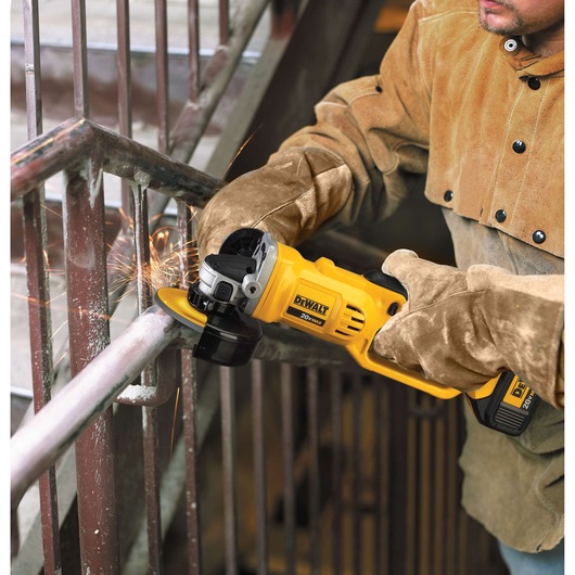 A person wearing protective gloves and a brown work jacket is using a DEWALT DCG412B cordless angle grinder to cut or grind a metal railing. Sparks are flying from the point of contact.