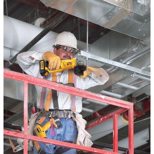 A worker wearing safety gear uses a DEWALT DCG412B cordless angle grinder to cut metal pipes on a construction site. The worker is standing on a red scaffold and is surrounded by industrial ductwork.