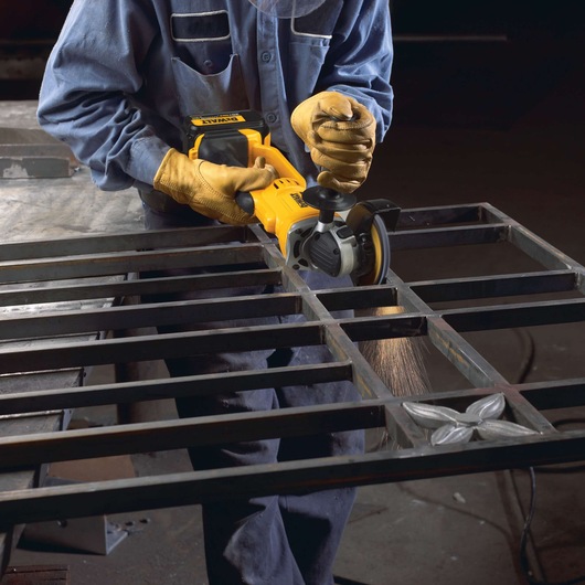 A person wearing protective gloves and a blue work uniform is using a DEWALT cordless angle grinder (model DCG412B) to grind or cut a metal grid structure. Sparks are visible from the contact point between the tool and the metal.