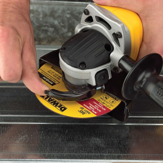 Close-up of a person adjusting the guard on a DEWALT angle grinder with a yellow and black grinding disc, positioned above a metallic surface.