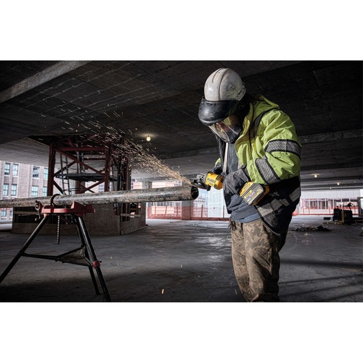 A worker wearing safety gear uses a DEWALT cordless grinder to cut a metal pipe in an industrial construction setting, with sparks flying from the grinding tool.