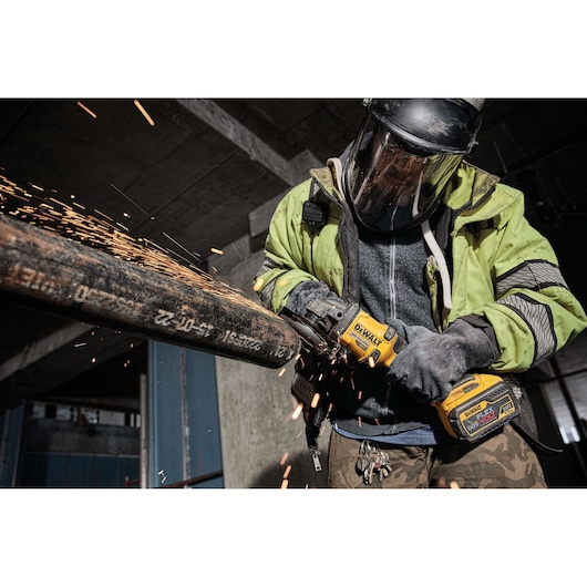 A person wearing safety gear uses a DEWALT cordless angle grinder to cut a metal pipe, producing sparks in an industrial environment.