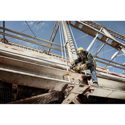 A worker wearing a yellow hard hat and safety gear uses a DEWALT power tool to cut through a metal beam on a large industrial bridge. Sparks are flying from the cutting area.