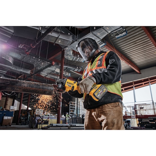 A person wearing safety gear uses a DEWALT cordless grinder to cut a metal pipe, creating sparks, inside a construction site.