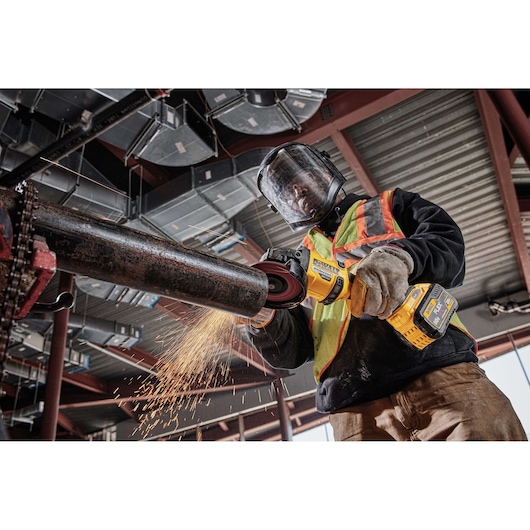 A worker wearing protective gear, including a face shield, gloves, and a high-visibility vest, using a DEWALT cordless angle grinder to cut a metal pipe, with sparks flying in an industrial setting.