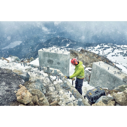 A worker wearing safety gear uses a tool near large concrete blocks on a snowy mountain construction site. There is a DEWALT tool case on the ground, and the landscape is mountainous with snow and rocks.