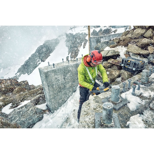 A worker wearing a helmet, ear protection, and a green jacket uses a DEWALT power tool on a snowy construction site in a mountainous area. Concrete structures and metal bolts are visible, along with a DEWALT tool case resting on rocks.