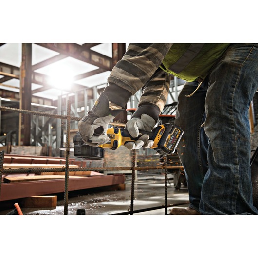 A person wearing gloves and work clothes uses a DEWALT cordless angle grinder to cut metal rods at a construction site, with sparks flying.