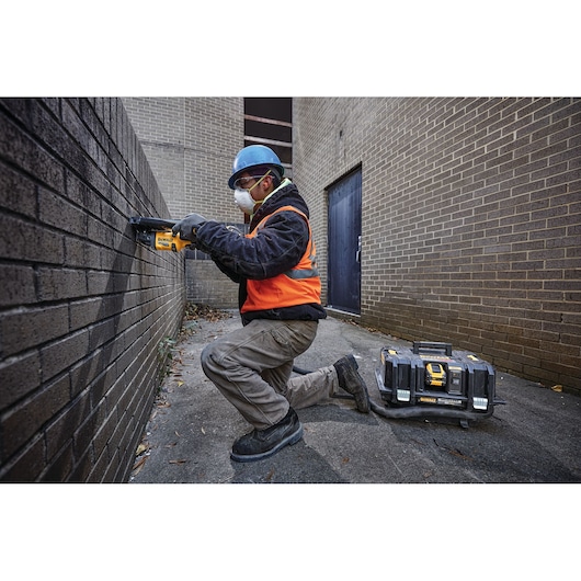 A worker wearing a blue hard hat, orange safety vest, and gloves is kneeling while using a DEWALT power tool on a brick wall in an outdoor alleyway. A DEWALT toolbox with a hose attachment is placed on the ground beside the worker.