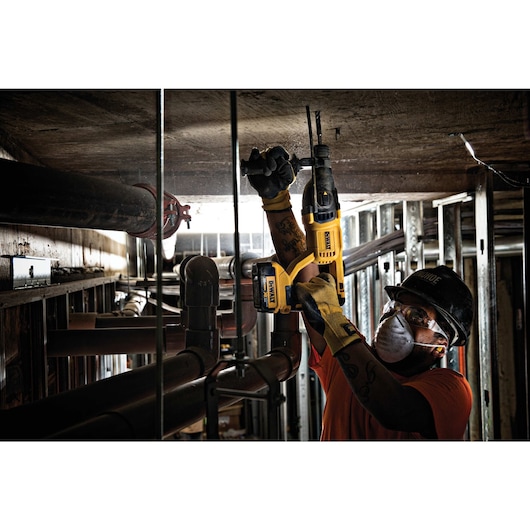 A person wearing gloves and a hard hat is using a yellow DEWALT cordless rotary hammer drill (model DCH133B) to drill into a concrete ceiling in an industrial environment with metal pipes and framing.