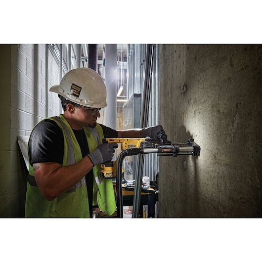 A construction worker wearing a hard hat and safety vest is using a DEWALT cordless rotary hammer drill (model DCH133B) with a dust extraction attachment to drill into a concrete wall inside an industrial building. The worker's face is blurred for privacy.