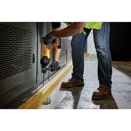 A person using a DEWALT cordless rotary hammer drill to drill into a metal surface near the floor. The person is wearing jeans, work boots, gloves, and a safety vest.