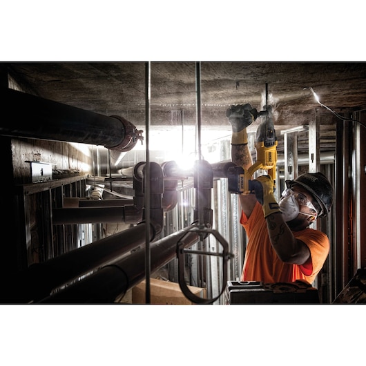 A construction worker uses a DEWALT cordless hammer drill to drill into a ceiling in an industrial setting. The worker is wearing an orange shirt, gloves, and a hard hat, surrounded by metal pipes and construction materials.
