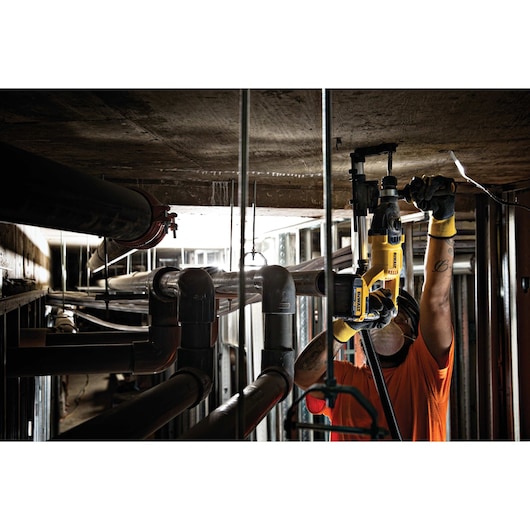 A worker in a construction area uses a yellow DEWALT power tool to drill into the ceiling surrounded by pipes.