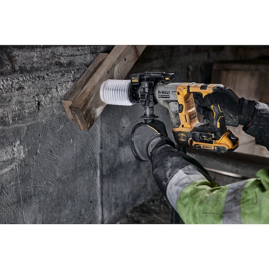 A person using a DEWALT cordless rotary hammer drill to bore a hole through a wooden beam and concrete wall. The user is wearing gloves and a work jacket.