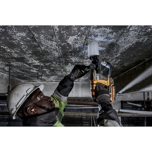 A person wearing safety gear and gloves is using a DEWALT cordless rotary hammer drill (model DCH172B) to drill into a concrete ceiling. The tool is equipped with a dust extraction attachment.
