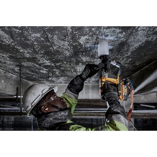 A worker wearing safety gear, including a helmet, gloves, and protective glasses, is using a yellow DEWALT power tool to drill into a concrete ceiling in an industrial setting.