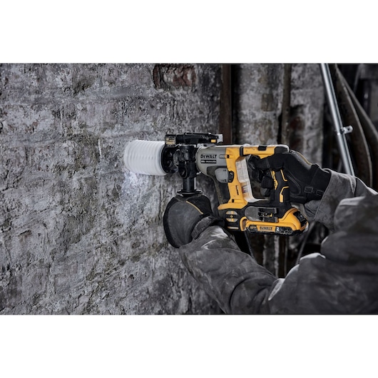 A person wearing protective gloves and clothing is using a yellow and black DEWALT cordless rotary hammer drill to create a hole in a concrete or brick wall.