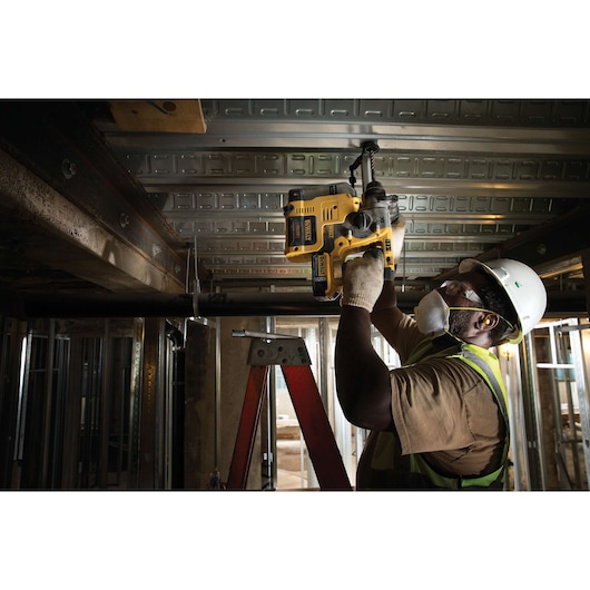 A construction worker wearing a hard hat, safety glasses, face mask, gloves, and a safety vest is using a yellow DEWALT cordless rotary hammer drill to work on a metal ceiling from a ladder inside a building under construction.