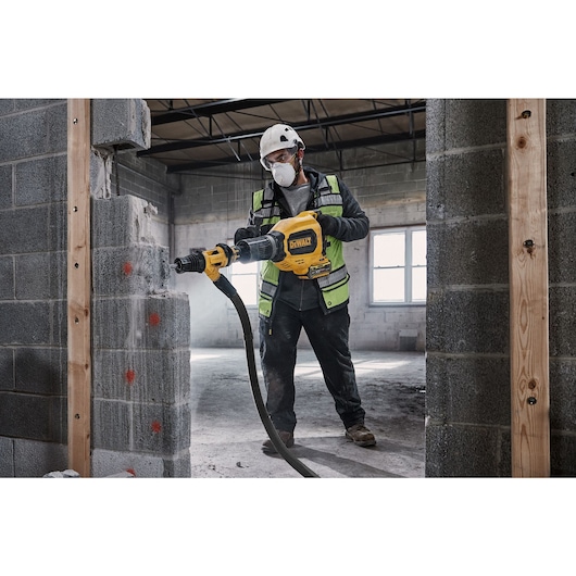 A person wearing a helmet and safety vest using a DEWALT cordless rotary hammer drill to break through a concrete block wall in an unfinished construction area.