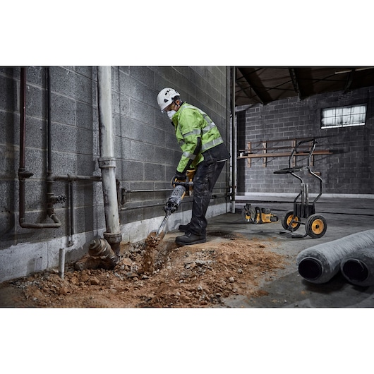 A person wearing safety gear uses a DEWALT demolition hammer to break up a concrete floor near a wall in an industrial setting. Construction tools and equipment are visible in the background.