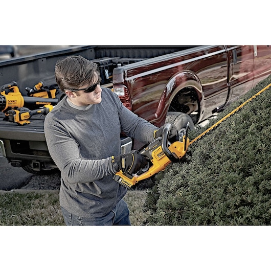A person wearing gloves is trimming a hedge with a yellow DEWALT cordless hedge trimmer (model DCHT820B) next to a pickup truck, with additional DEWALT tools visible in the truck bed.