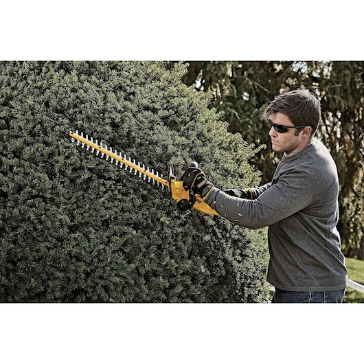 A person using a DEWALT cordless hedge trimmer to trim a large bush outdoors. The person is wearing gloves and a long-sleeve shirt for protection.