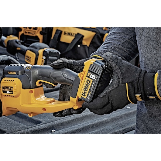A close-up of a person wearing gloves and inserting a DEWALT 20V MAX XR lithium ion battery into a yellow and black cordless power tool. Other DEWALT tools and bags are visible in the background.
