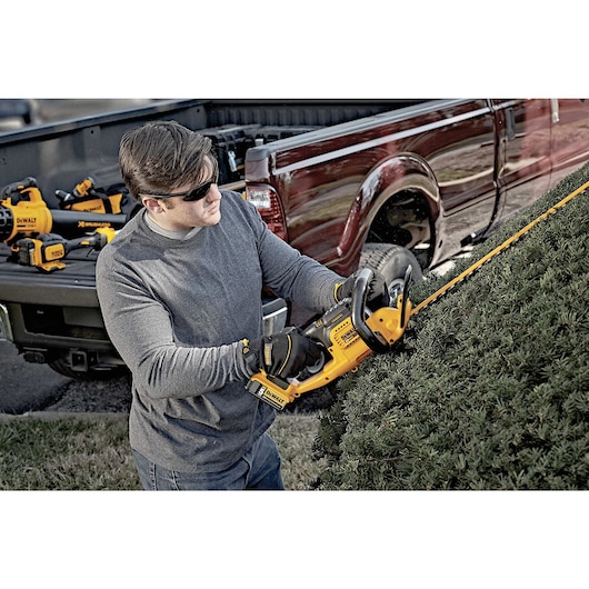 A person using a DEWALT cordless hedge trimmer to trim bushes outdoors, with a pickup truck in the background containing other DEWALT power tools. The individual is wearing gloves and a long-sleeve shirt for protection.