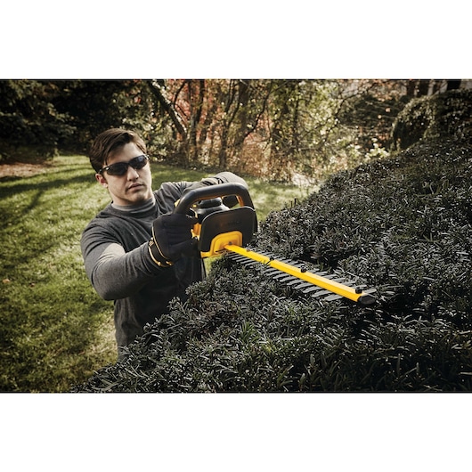 A person is using a DEWALT cordless hedge trimmer outdoors to trim bushes. The tool is yellow and black, and the person's face is blurred.