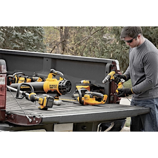 A person with a blurred face is standing next to a pickup truck with the tailgate down, organizing several yellow and black DEWALT power tools, including a hedge trimmer, a blower, a drill, and other cordless tools, in the truck bed.