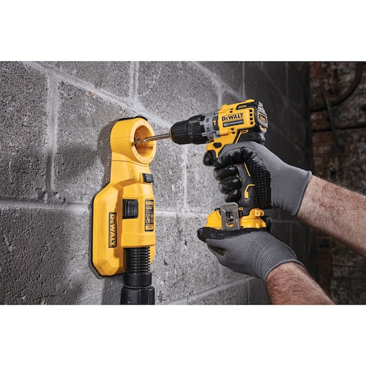 A person wearing work gloves uses a DEWALT cordless drill to drill into a concrete wall. The drill is attached to a yellow dust extraction device mounted on the wall, helping collect debris as the hole is made.