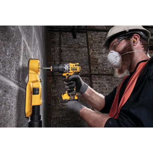 A person wearing safety gear uses a yellow DEWALT cordless drill to drill into a concrete wall. The person is also using a yellow attachment connected to a vacuum for dust extraction.
