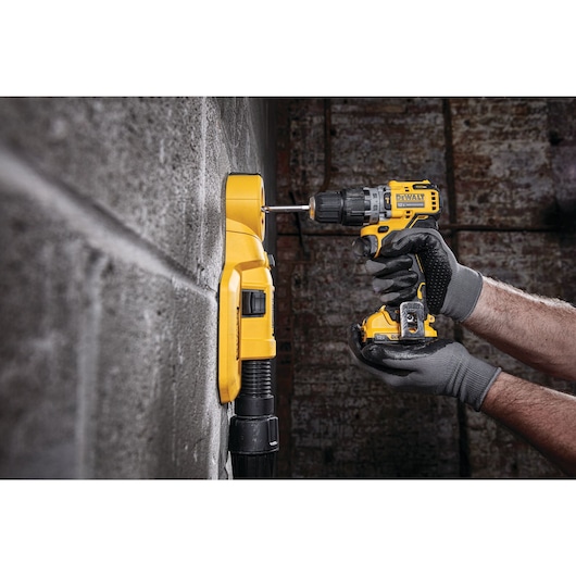 A person wearing gray gloves is using a yellow DEWALT cordless drill to drill into a concrete wall. The drill is being used alongside a yellow wall-mounted dust collector, which helps capture debris during the drilling process.