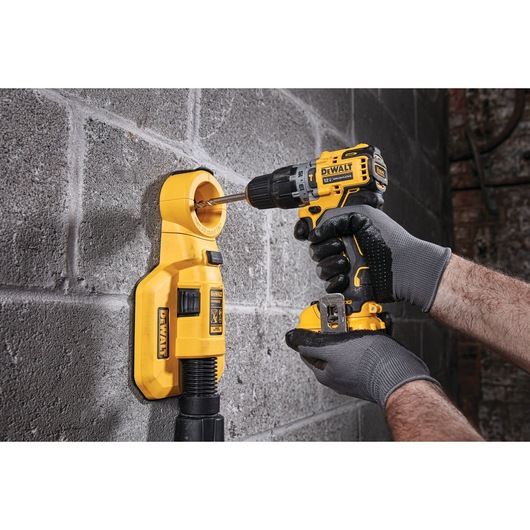 A person wearing gloves uses a DEWALT cordless drill to drill into a concrete wall. The drill is attached to a yellow DEWALT dust extractor that collects dust while drilling.