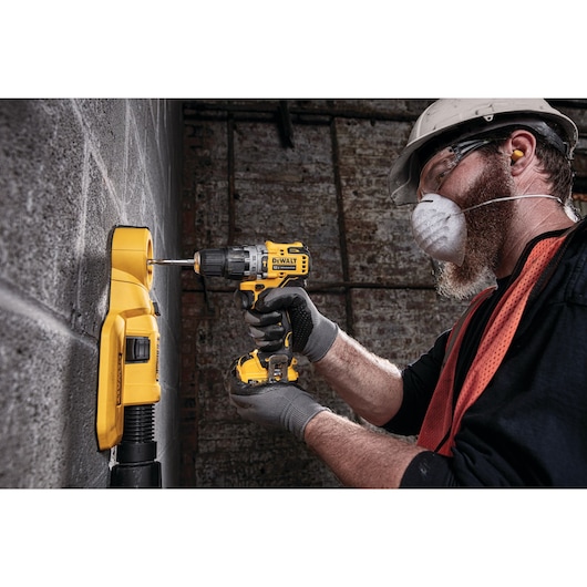 A person wearing protective gear uses a DEWALT cordless drill to drill into a concrete wall, with a yellow dust extraction tool attached.