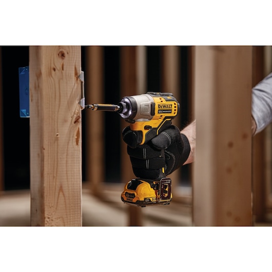 A person wearing black gloves uses a yellow and black DEWALT cordless impact driver to drive a screw into a wooden frame.