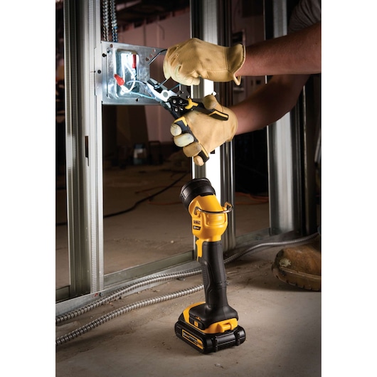 A DEWALT cordless LED work light (model DCL040) is placed on the floor and illuminating an electrical wiring installation. A person wearing gloves is using pliers to work on wires inside a metal electrical box, with the light aiding visibility in a construction setting.