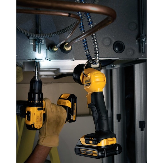 A DEWALT DCL040 handheld work light is hanging from a metal fixture, illuminating a workspace where someone wearing gloves is using a DEWALT cordless drill on an overhead surface with electrical and copper wiring visible.