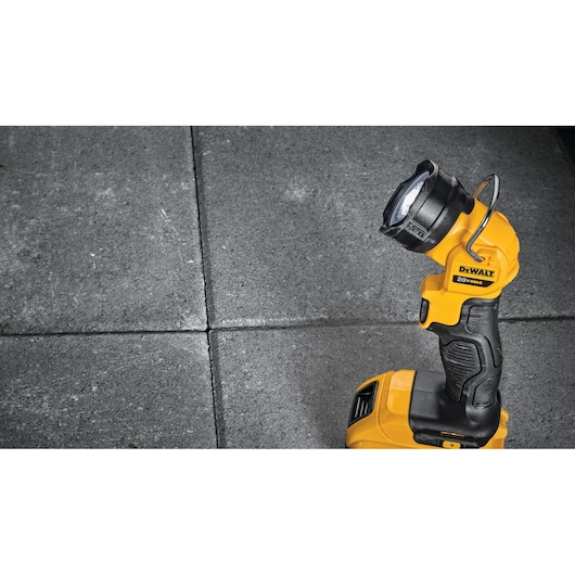 A DEWALT DCL040 20V handheld LED work light is placed on a gray tiled surface. The light is yellow and black with a handle and a metal hook for hanging.
