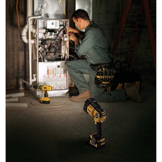 A mechanic is working on a furnace in a dimly lit room. In the foreground is a DEWALT DCL043 cordless spotlight, and next to the furnace is a yellow DEWALT cordless drill. The worker is wearing a green uniform and tan work boots, with DEWALT tools and a tool pouch nearby.
