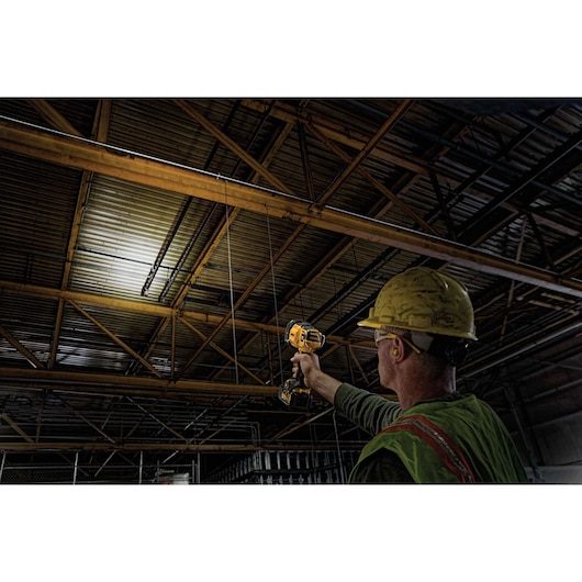 A person wearing a yellow hard hat and safety vest uses a DEWALT handheld spotlight to illuminate a metal ceiling inside an industrial building.