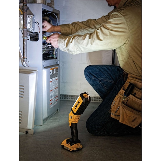 A person is repairing or inspecting an open furnace or electrical unit. A DEWALT DCL050 cordless work light is standing upright on the floor, illuminating the workspace. The person is wearing a tool belt and work clothes.
