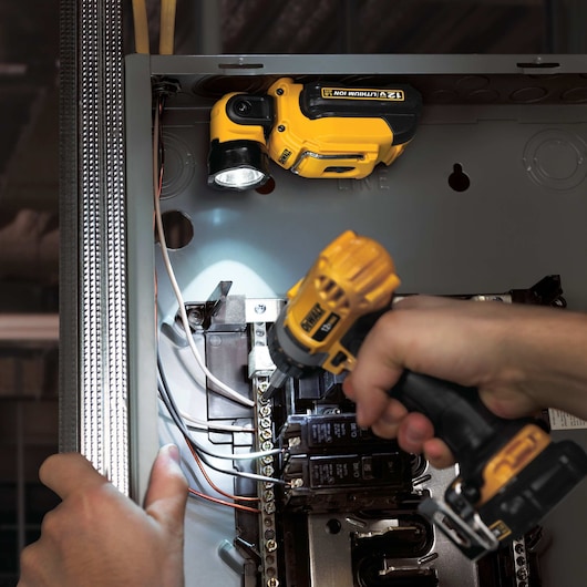 DEWALT portable LED flashlight illuminates an electrical panel while a person uses a DEWALT cordless drill to fasten wires inside the panel.
