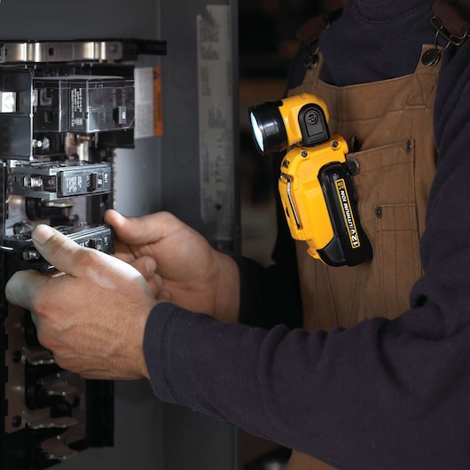 A person wearing brown overalls uses an inspection light attached to their chest while working inside an electrical panel. The portable yellow DEWALT light illuminates the area, aiding visibility during maintenance.