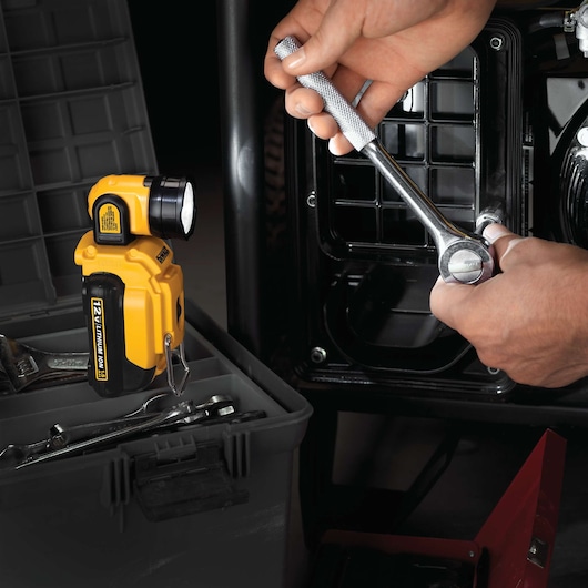 A DEWALT DCL510 12V Lithium Ion work light is positioned next to an open toolbox containing wrenches. Nearby, a person is using a ratchet to work on a metal surface or machinery part, illuminated by the work light.